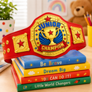 A red and gold 'Junior Champion' toy belt is displayed across a stack of colorful children's books on a light wooden table. The books read 'Be Brave', 'Dream Big', 'YOU CAN DO it!', and 'Little World Changers'.