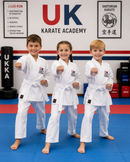Three children in white karate uniforms posing in a martial arts studio with 'UK Karate Academy' branding.
